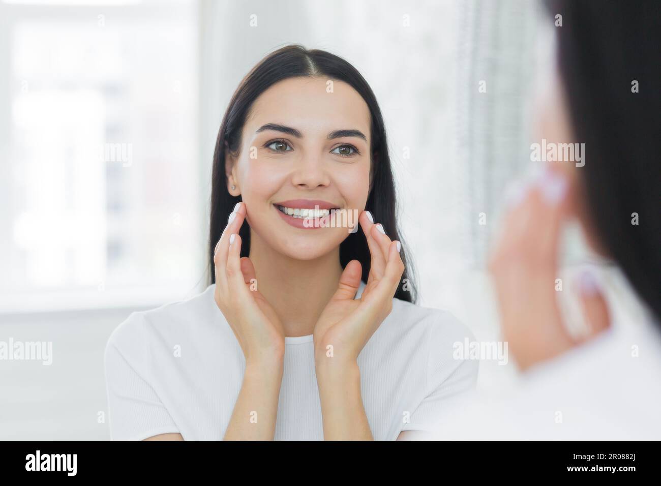 Close-up photo. A young woman stands in front of a mirror at home and looks at her reflection. She puts cream on her face, makes up, massages with her hands, smiles. Stock Photo