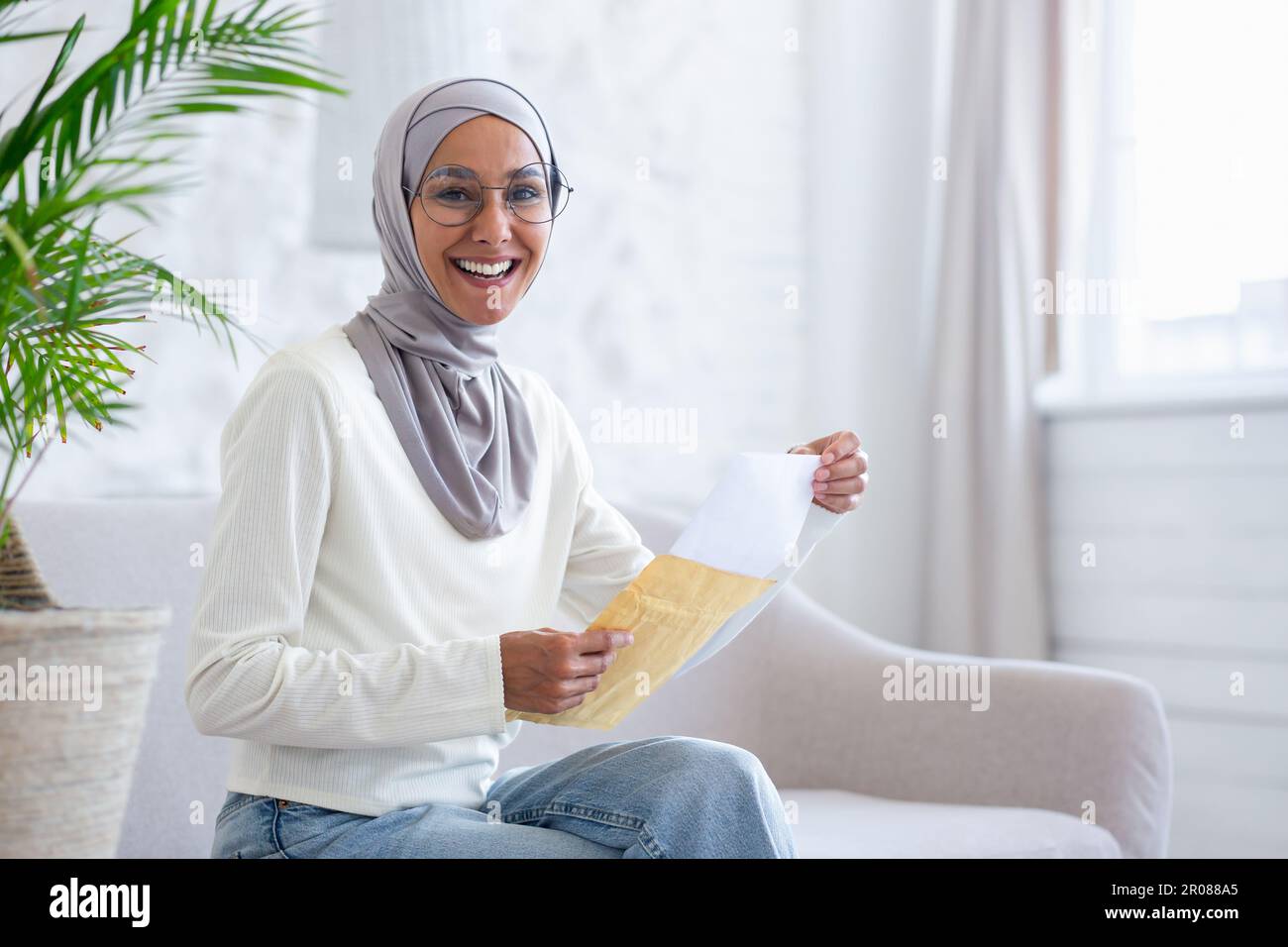Happy young muslim woman in hijab sitting on sofa at home and opening ...