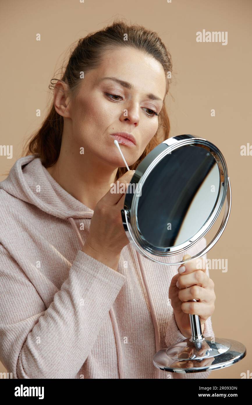 female with herpes on lips and mirror applying ointment using cotton swab isolated on beige
