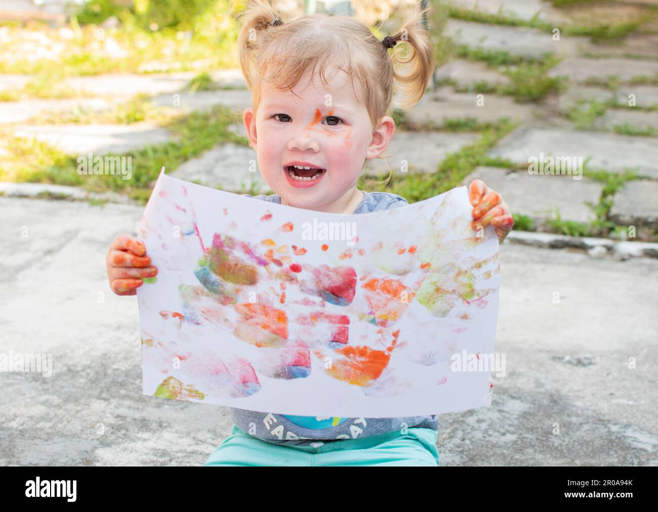 Happy Caucasian toddler girl with 2 pigtails showing off her artwork made with finger paints. Sensory art activities for kids. Stock Photo