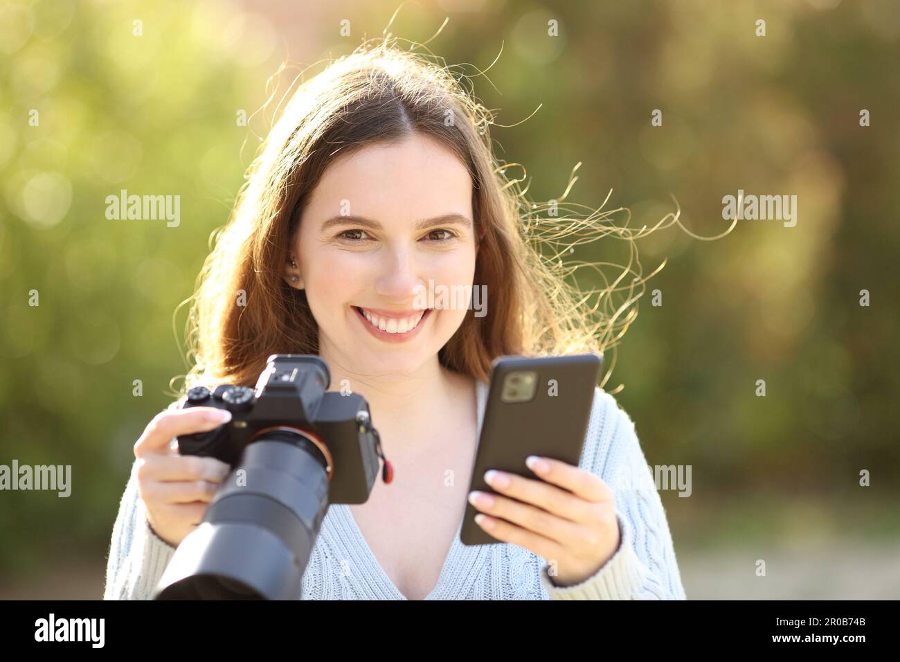 Happy photographer looks at you holding cell phone and mirrorless camera in a park Stock Photo