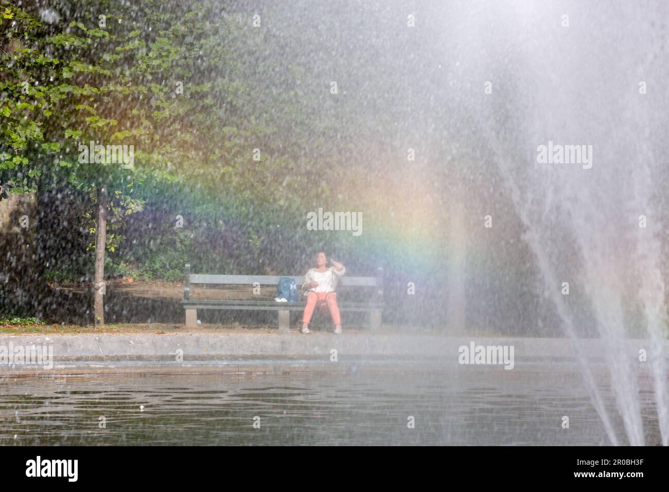 Lady seated on a bench in the Parc de Bruxelles, seen through a fountain with rainbow effect. Stock Photo