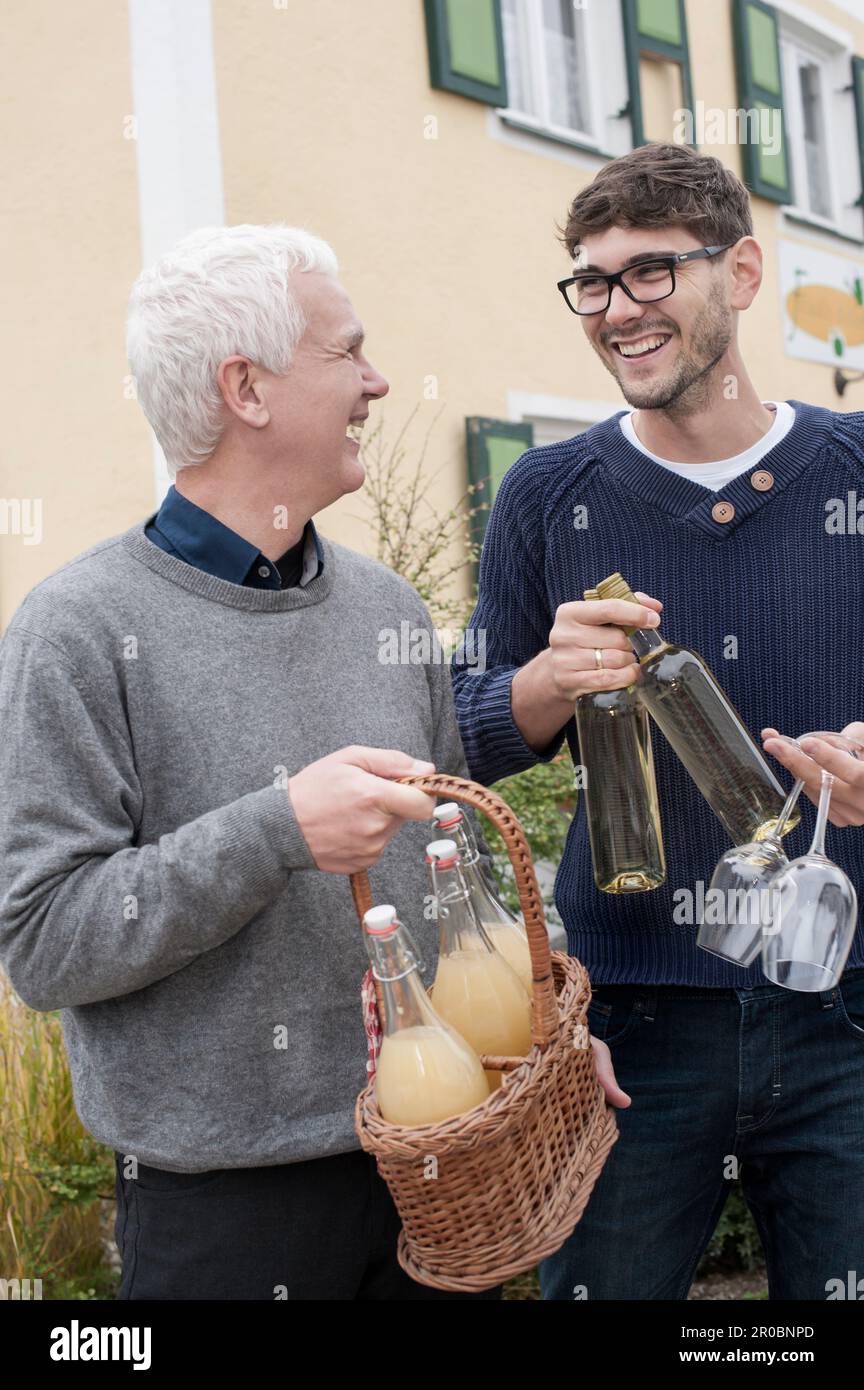 Father and son holding bottles of white wine and apple juices and laughing, Bavaria, Germany Stock Photo