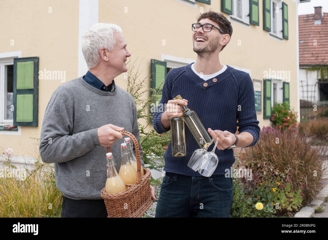 Father and son holding bottles of white wine and apple juices and laughing, Bavaria, Germany Stock Photo
