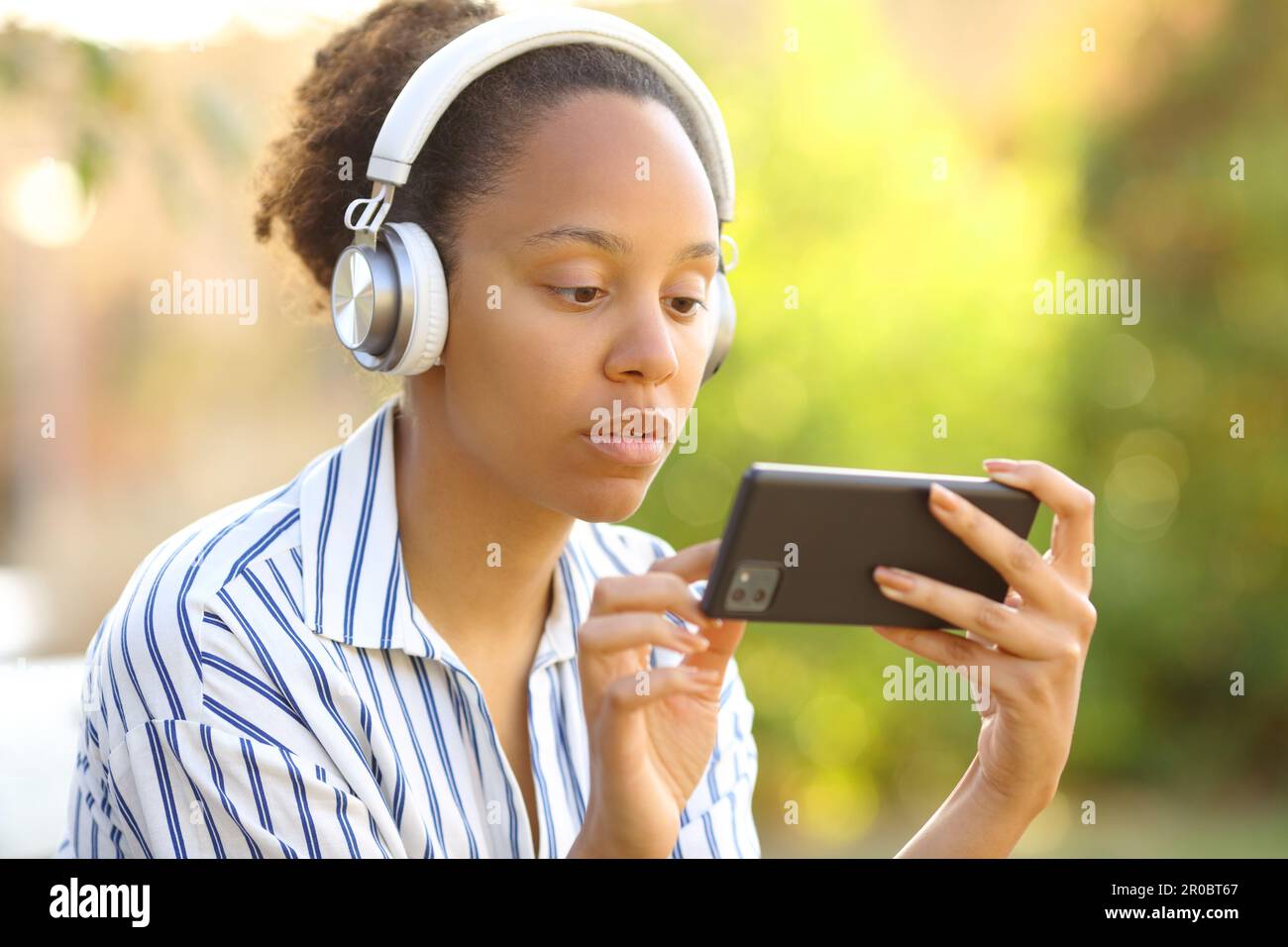 Serious black woman wearing headphone watching videos on phone in a park Stock Photo