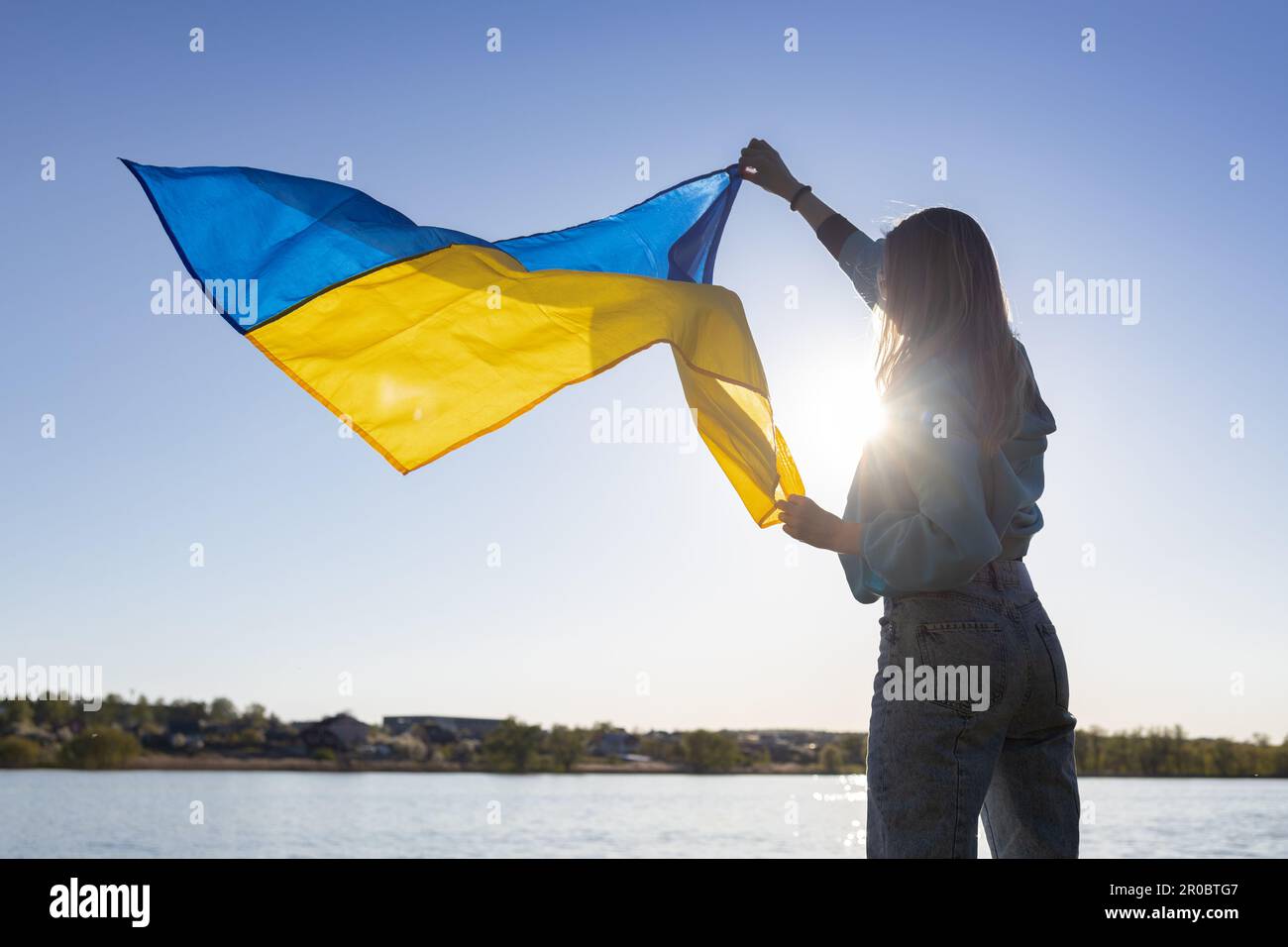 female silhouette holds a Ukrainian flag fluttering in the wind, beautifully illuminated by the sun Stock Photo