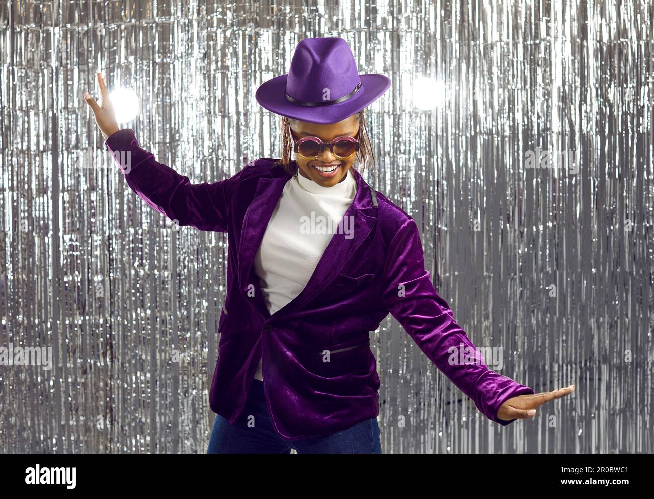 Happy confident young black woman in a purple hat, jacket and glasses dancing at a party Stock Photo