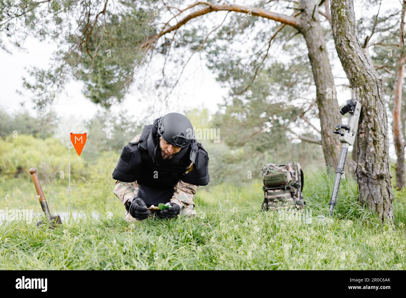 A man in a military uniform and a bulletproof vest works in the forest to demine the territory. A man warns of danger by making a red mark. Stock Photo