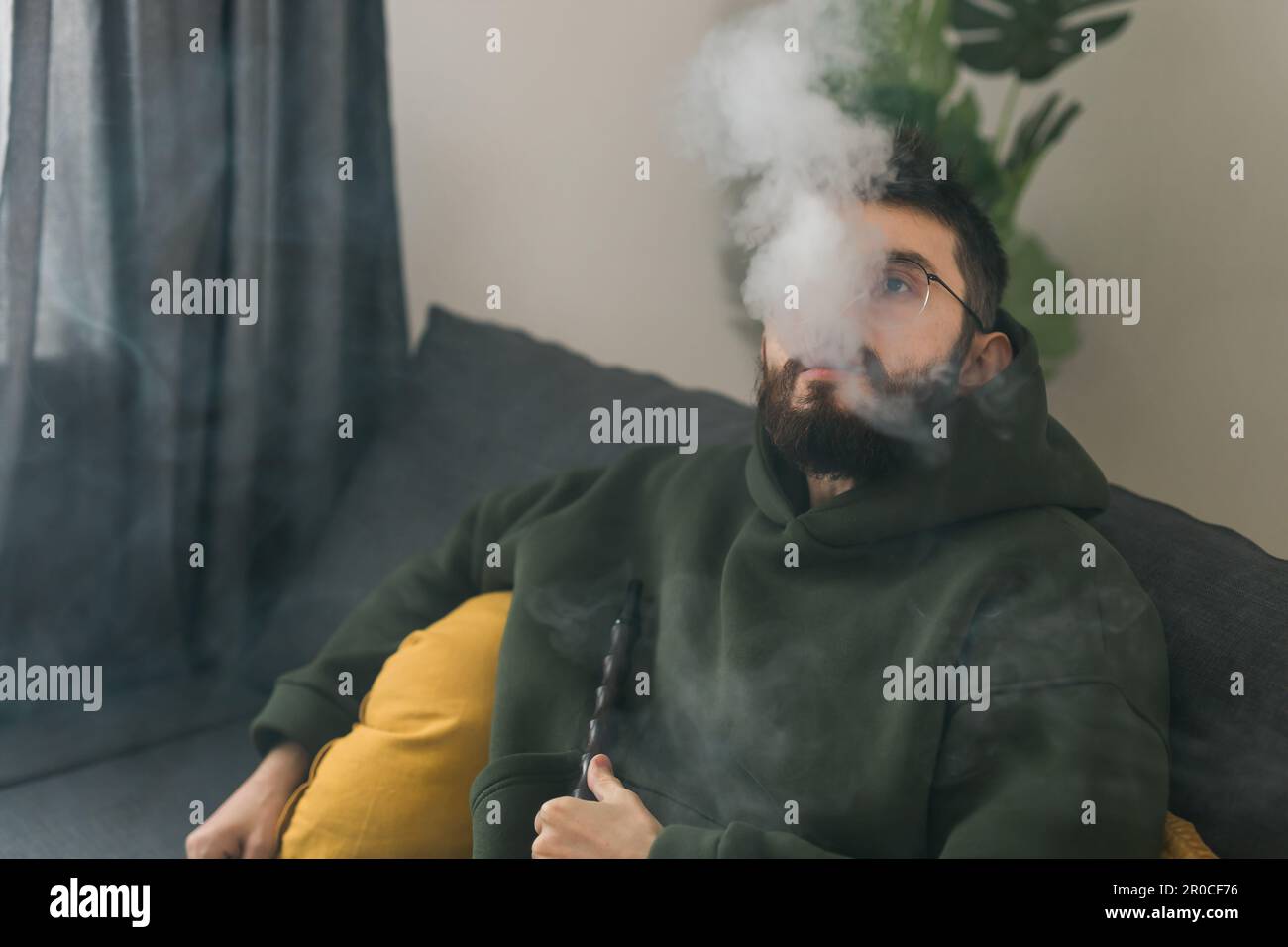 Bearded millennial or gen z man smoking hookah while relaxing on sofa