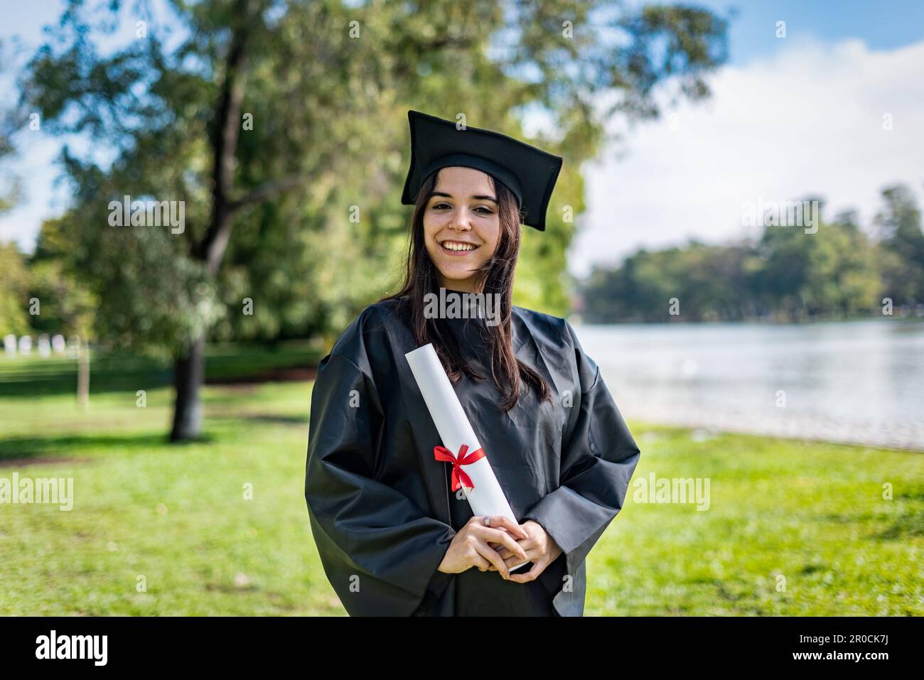 Happy caucasian graduated girl with long brown hair. She is wearing a bachelor gown and a black mortarboard. She is holding a diploma Stock Photo