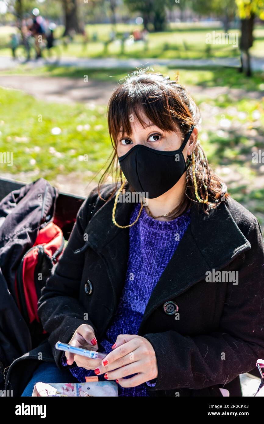 A woman with her mask on is reading a text message on her mobile phone in a public park Stock Photo