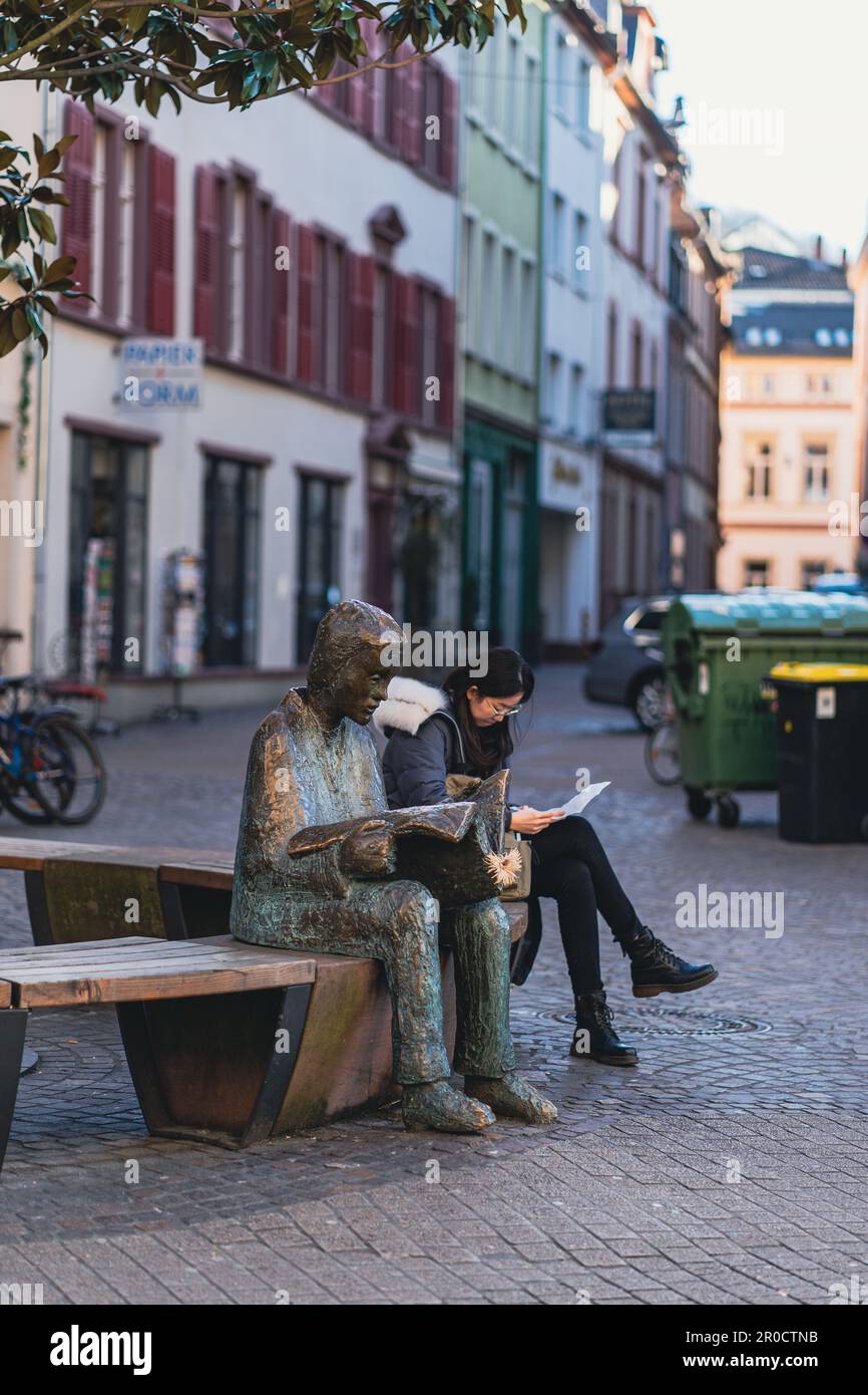 Bronze statue of a man reading newspaper. A young woman is sitting on a bench next to the statue and reading, Heidelberg, Germany Stock Photo