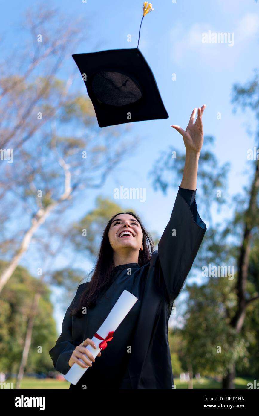 Happy caucasian graduated girl in a black gown throwing the mortarboard to the sky Stock Photo