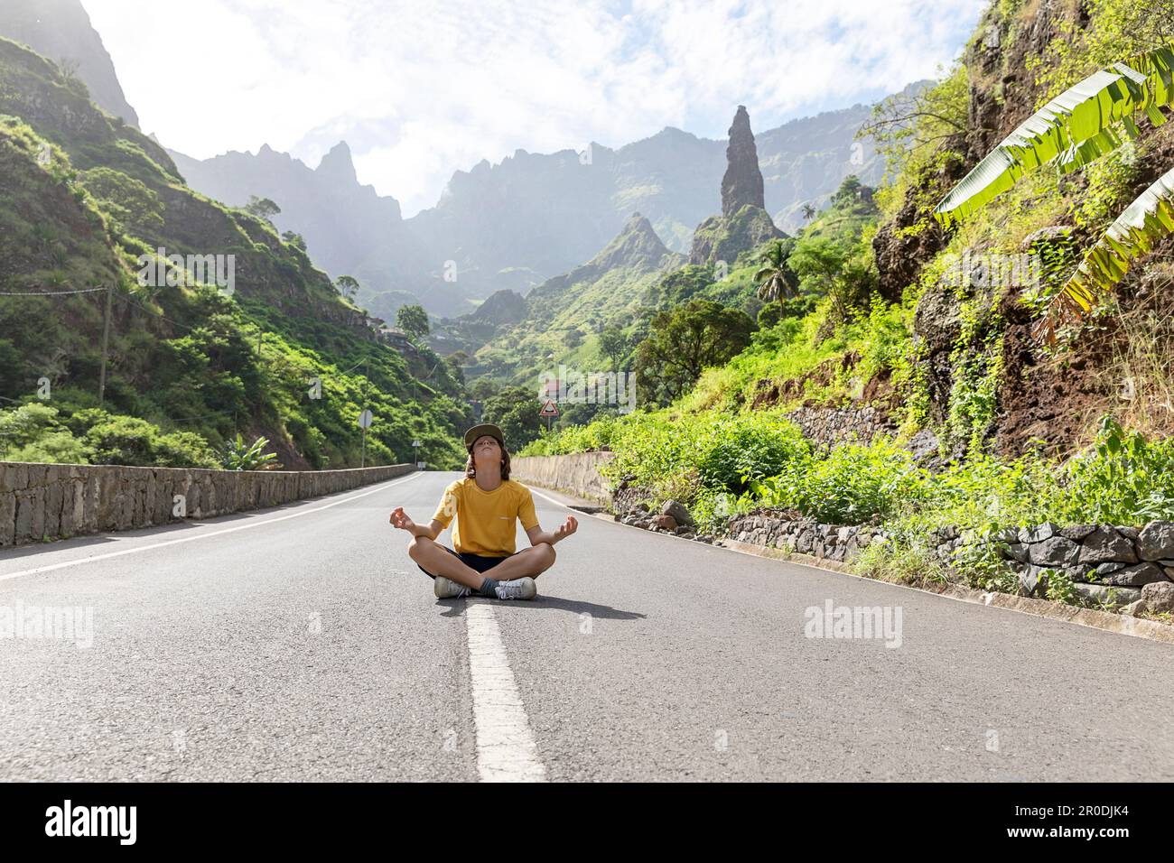 Tourist boy sitting in yoga position on a road in Xoxo village wit rock pillar in the background, lush valley on the island of Santo Antao, Capo verde Stock Photo