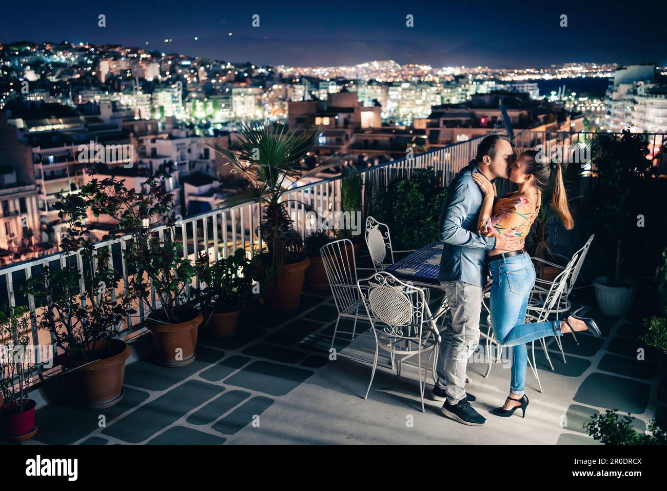 Couple at night, city lights view. Romantic date or proposal. Man and woman kiss and embrace on balcony or roof terrace. Dark blue sky. Stock Photo