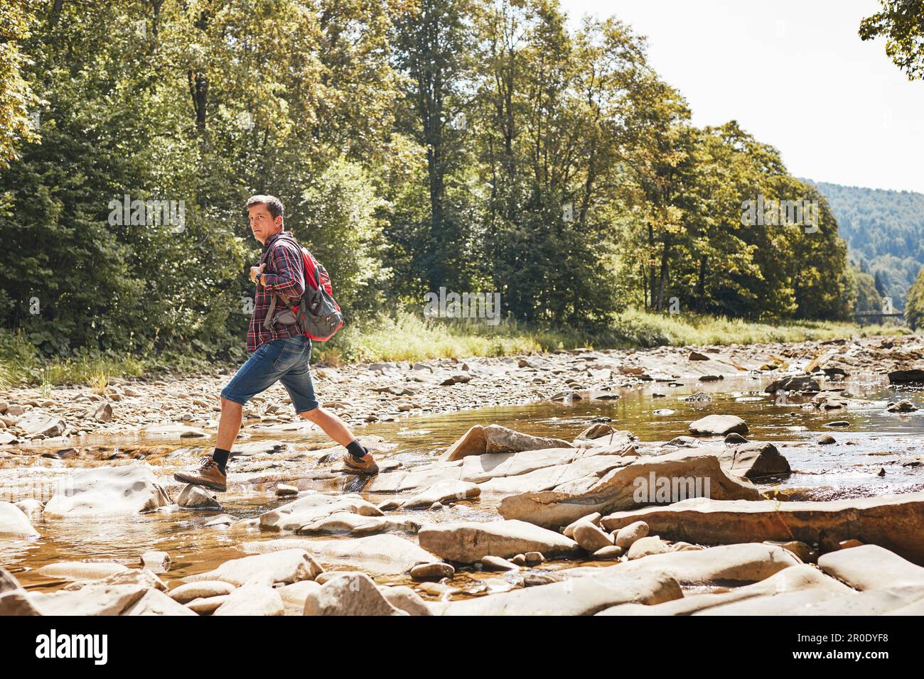 Trekking with backpack concept image. Backpacker wearing trekking boots crossing mountain river. Man hiking in mountains during summer trip. Vacation Stock Photo