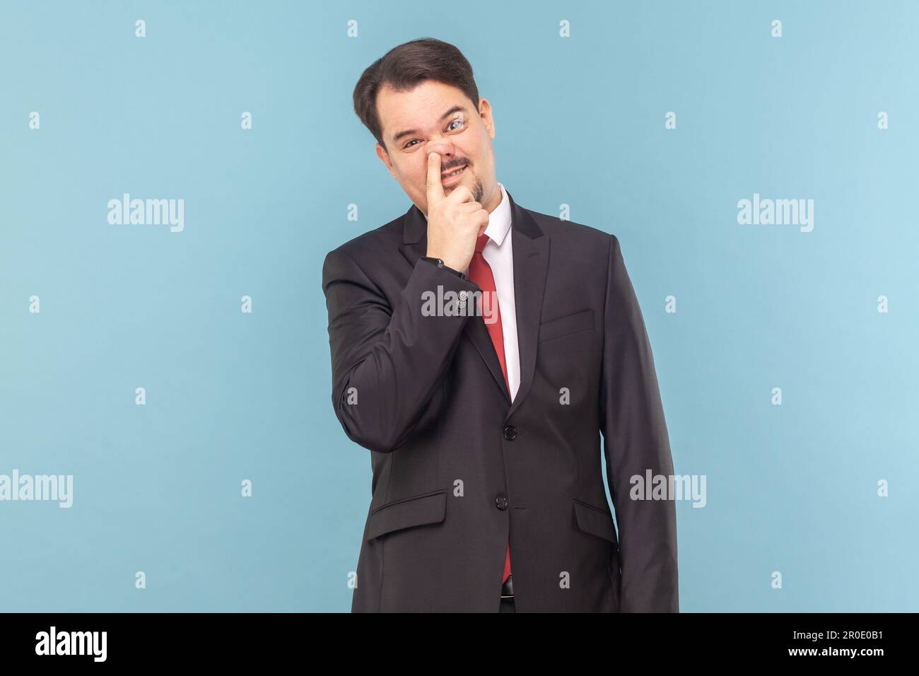 Portrait of funny childish man with mustache standing having comical facial expression, keeps finger in nose, wearing black suit with red tie. Indoor studio shot isolated on light blue background. Stock Photo