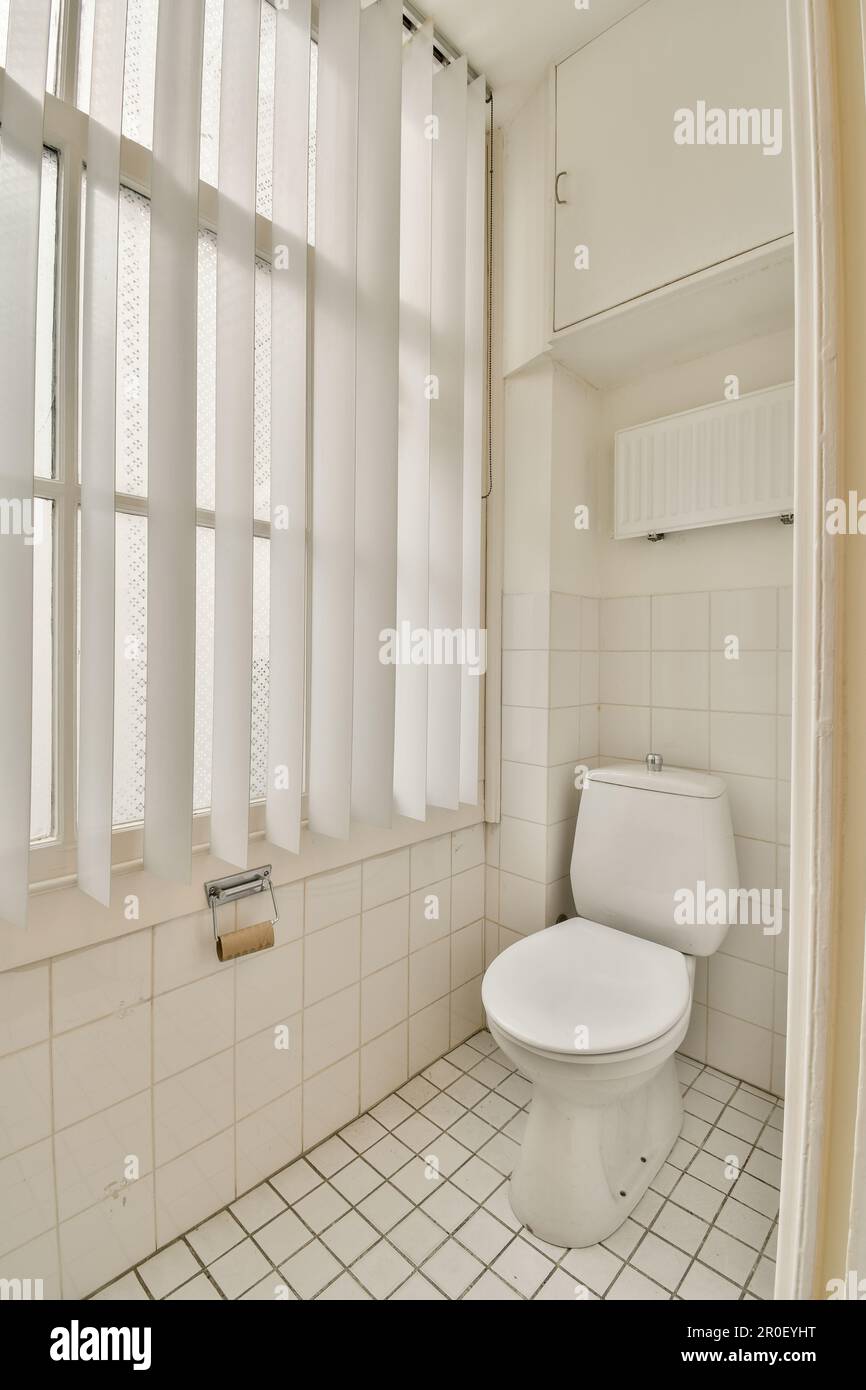a white toilet in a bathroom with windows on the wall and tiled floor