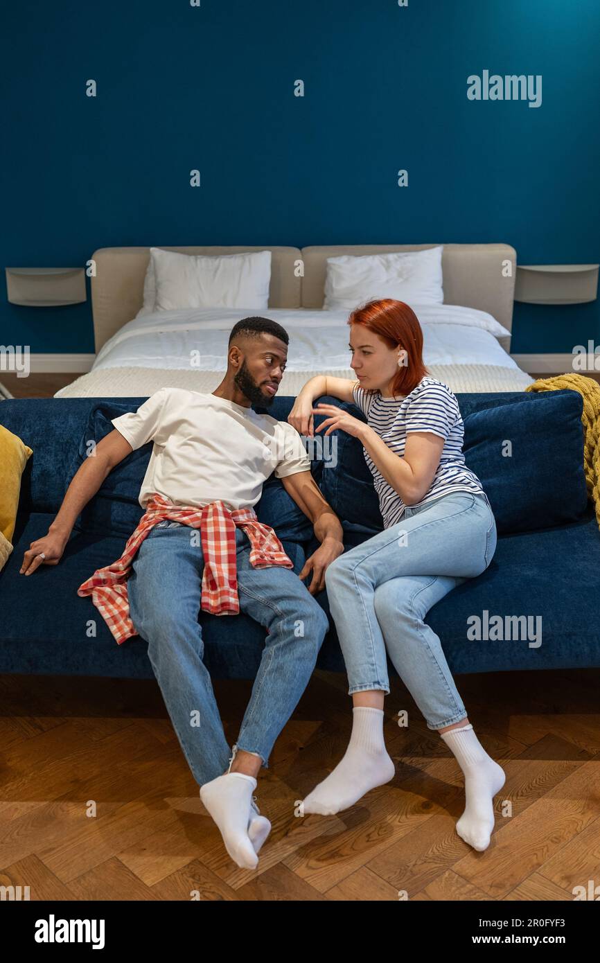 Conflicting multi ethnic stressed couple sitting on couch discussing talking about problems. Stock Photo