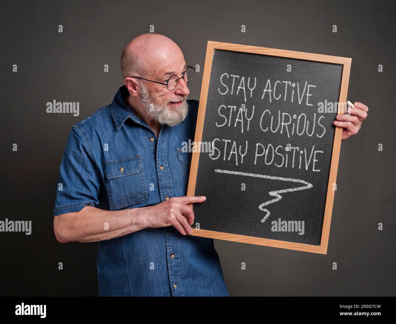 stay active, curious and positive - the keys to healthy aging, inspirational white chalk writing on a blackboard held by a senior man Stock Photo