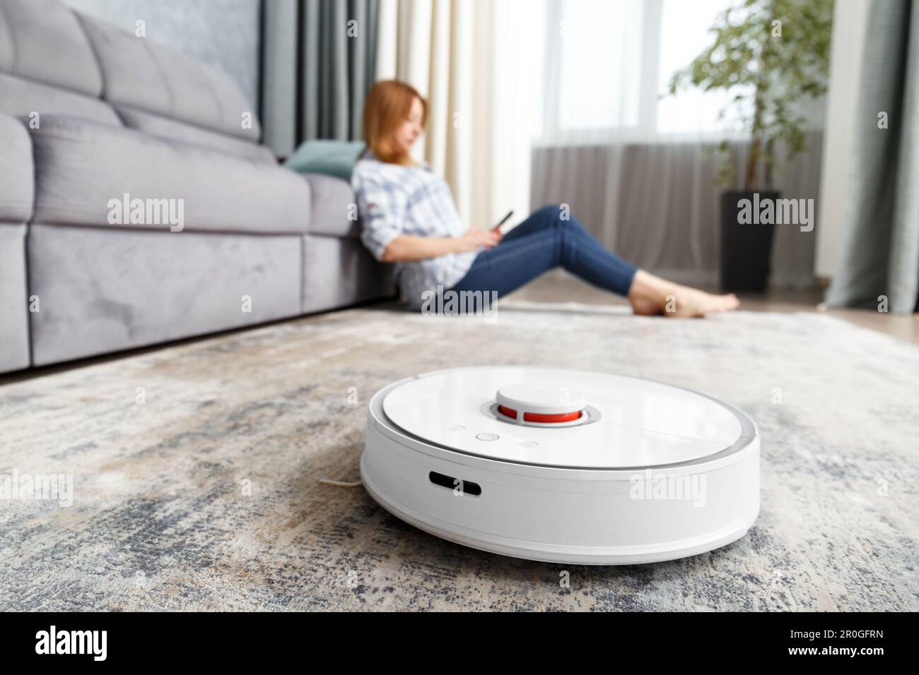 The girl is sitting in the living room and controls the robot vacuum cleaner using a smartphone. Smart House. The concept of smart home appliances Stock Photo