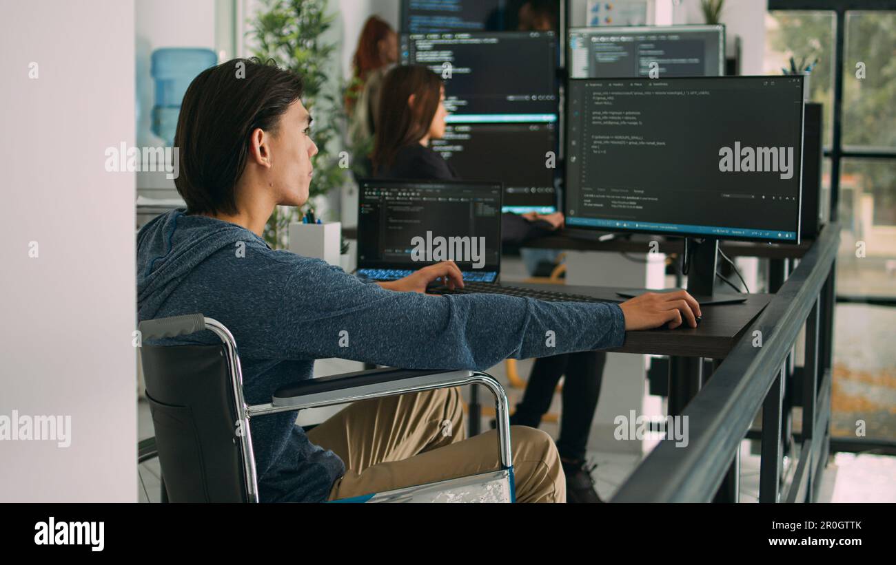 Focused software developer in wheelchair writing server code in agency data room, using security system. Engineer with physical disability programming user interface artificial intelligence office. Stock Photo