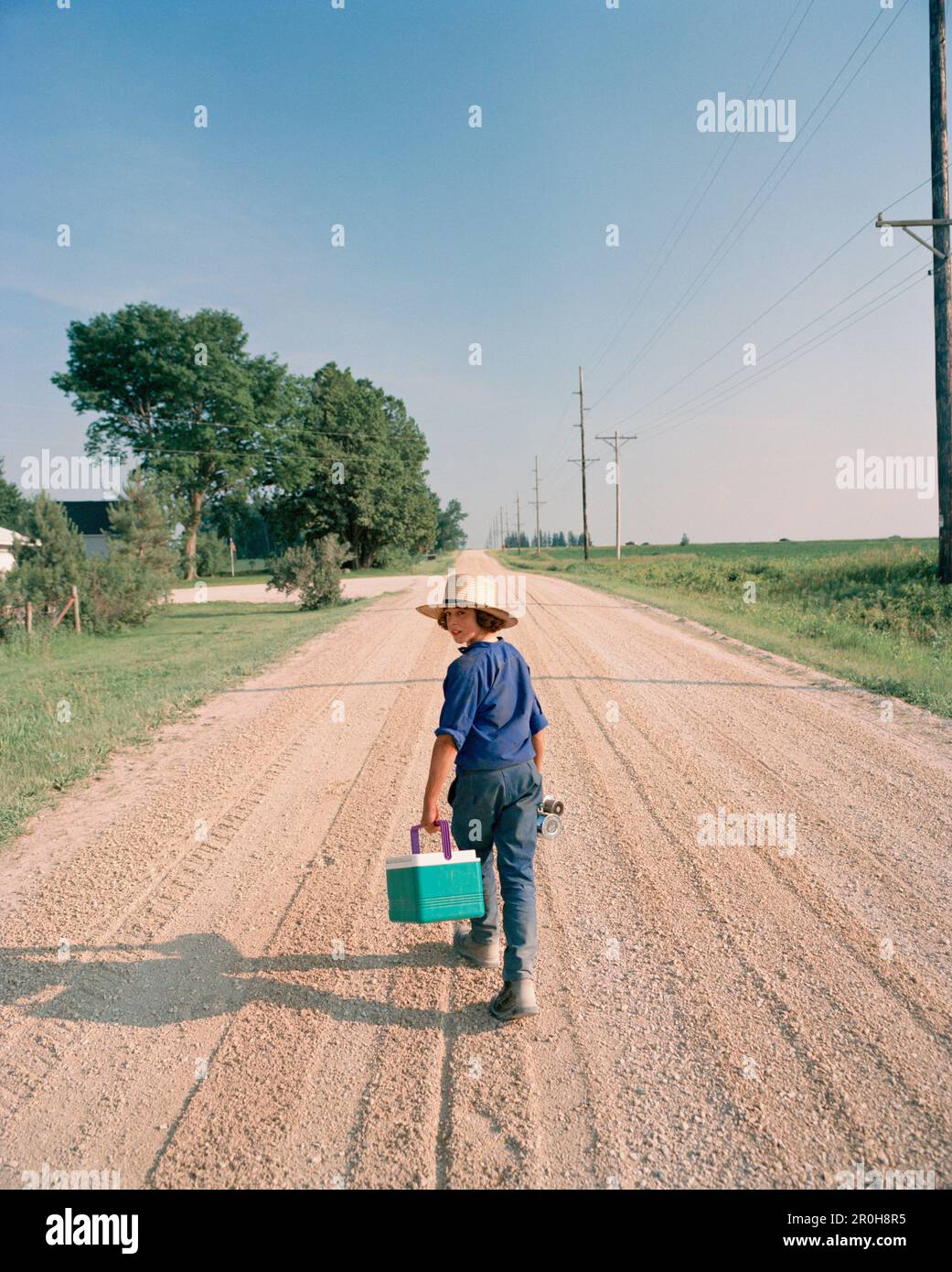 USA, Minessota, rear view of a young Amish boy walking on dirt road and carrying cooler Stock Photo