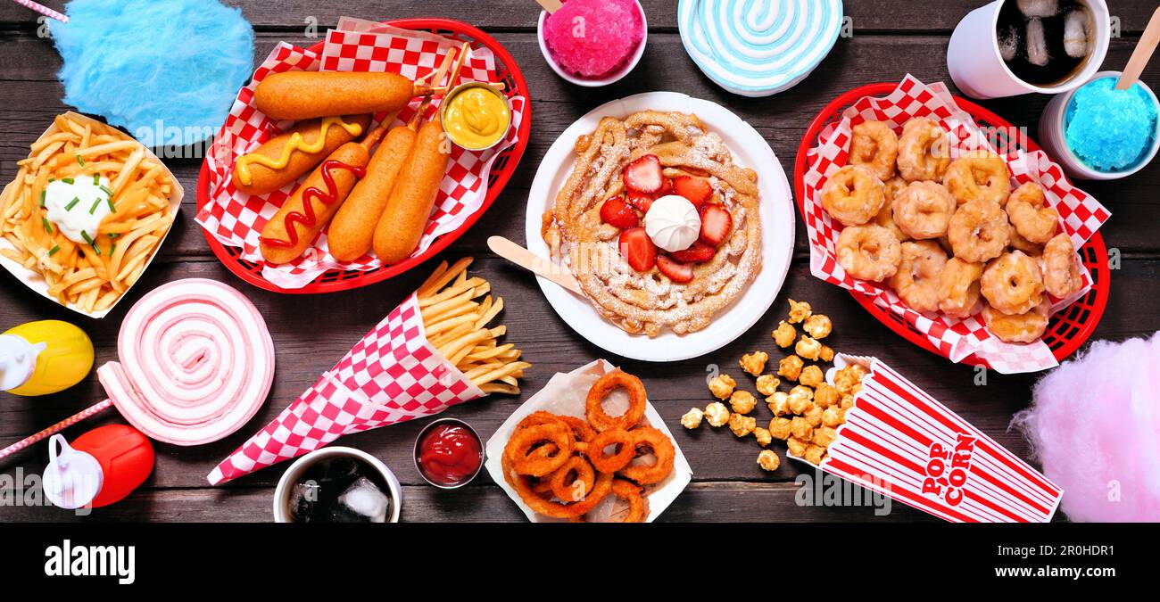 Carnival theme food table scene over a dark wood banner background. Top down view. Summer fair concept. Corn dogs, funnel cake, cotton candy and snack Stock Photo