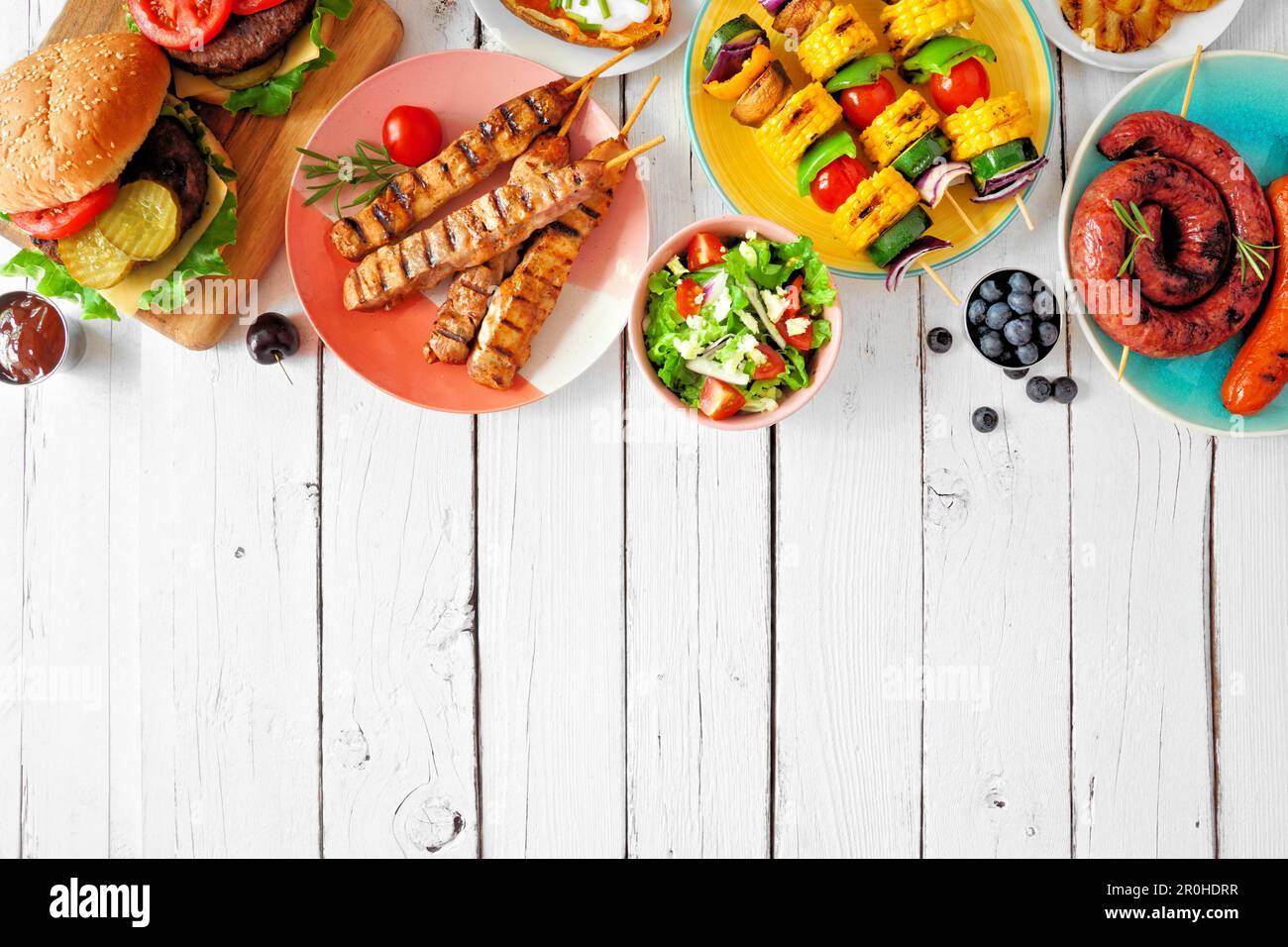 Summer BBQ or picnic food top border. Variety of burgers, grilled meat, vegetables, fruits, salad and potatoes. Overhead view on a white wood backgrou Stock Photo