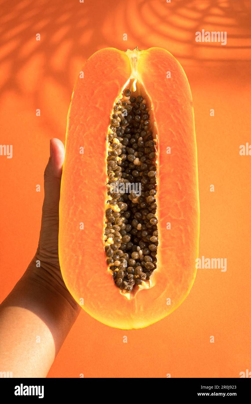 Hand holding papaya cut into half. On orange with palm leave shadow background. Summertime concept. Stock Photo