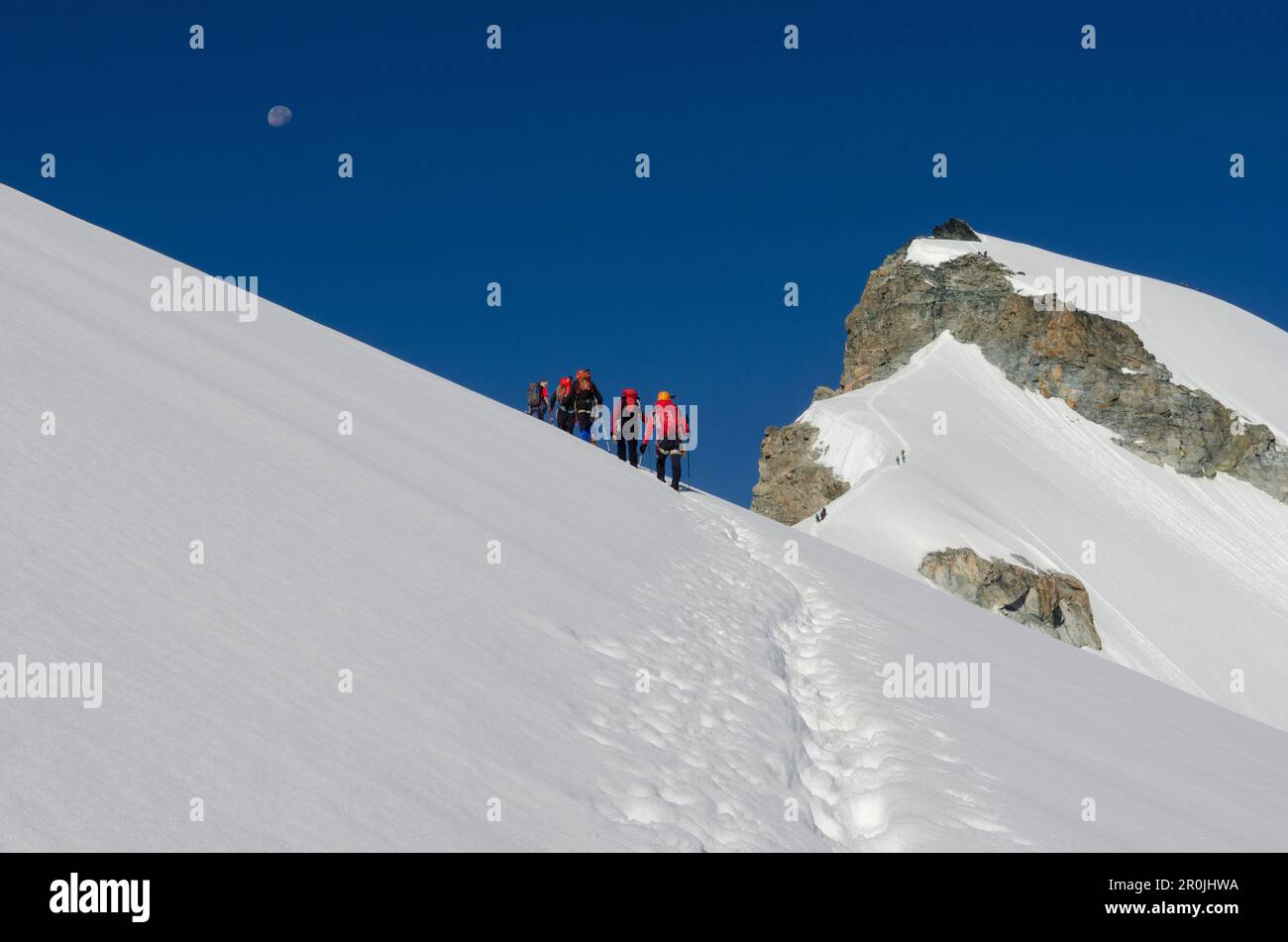 A group of alpinists on the snow crest of Hohlaub Ridge ascending towards the summit of Allalinhorn, above them the moon, Pennine Alps, canton of Vala Stock Photo