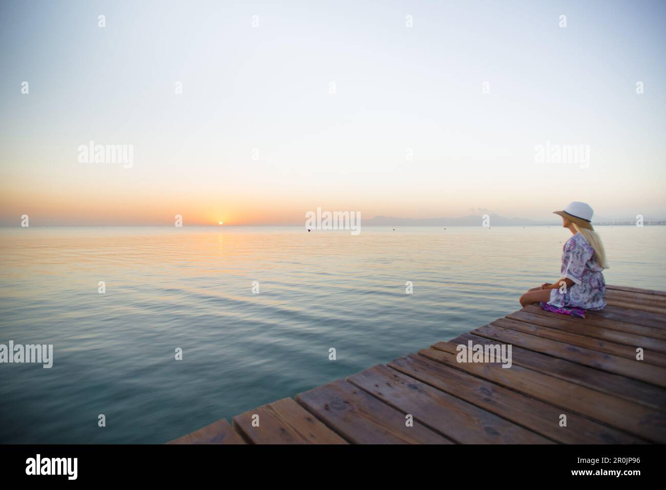 Young blond-haired woman sitting on the end of a long pier in the morning mood, enjoying the view of the sunrise. Playa de Muro beach, Alcudia, Mallor Stock Photo