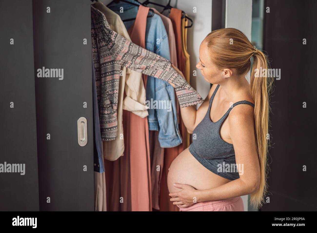 A Pregnant Woman Has Nothing To Wear. A Pregnant Woman Stands In Front Of A  Closet With Clothes And Does Not Know What To Wear Because The Clothes Do  Not Fit On
