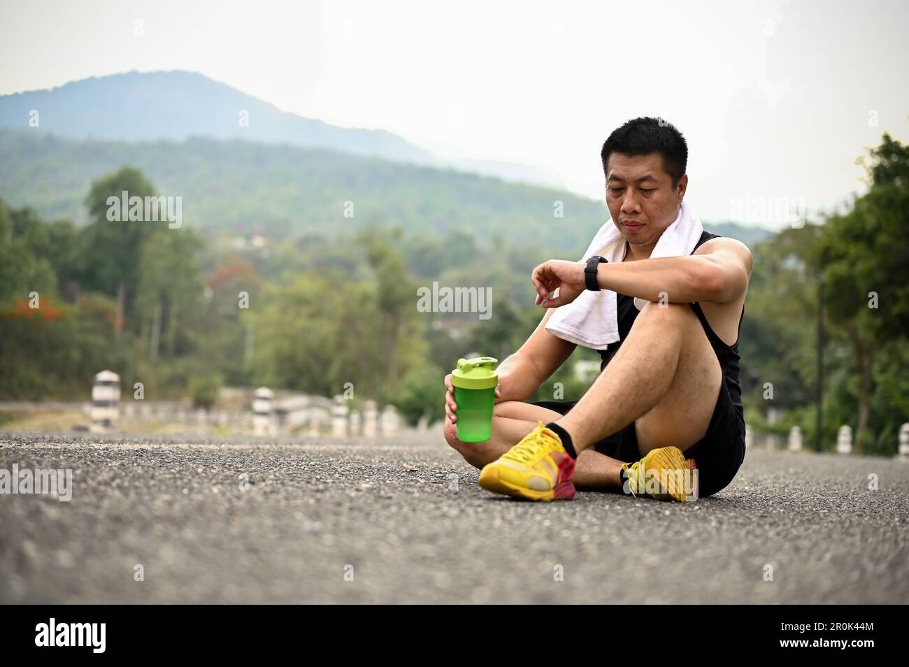 Tired and thirsty mature Asian man in sportswear sits on the road in a ...
