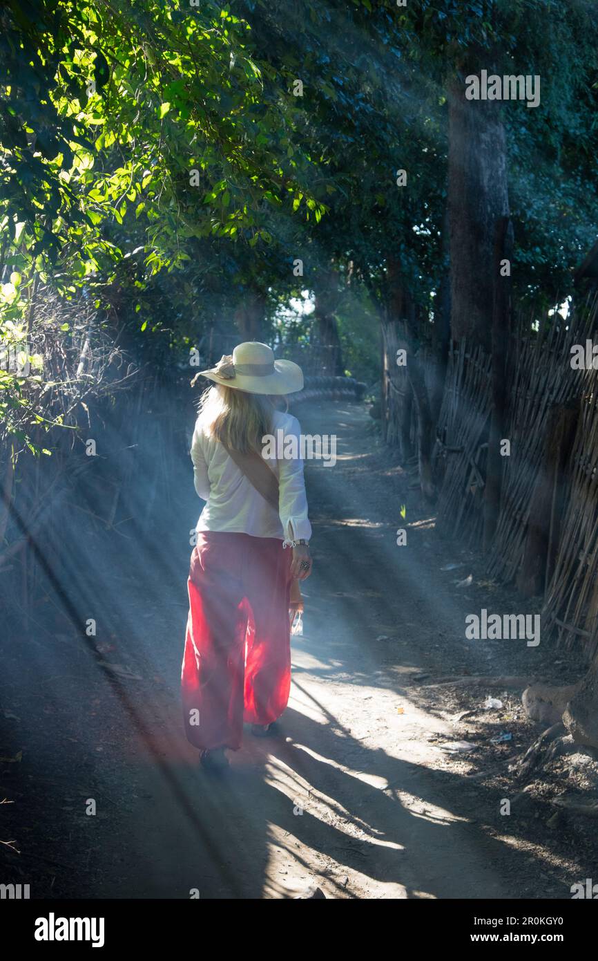 Young woman with sun hat strolls along path with sun rays shining ...
