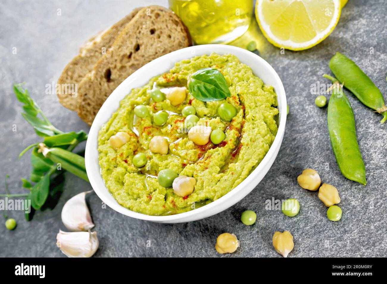 Hummus from green peas and chickpeas in a bowl, pea pods, lemon, garlic, vegetable oil in a decanter and slices of bread on granite countertop backgro Stock Photo