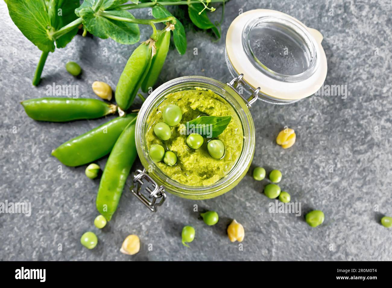 Hummus from green peas and chickpeas in a glass jar, pea pods on top of granite countertop background Stock Photo