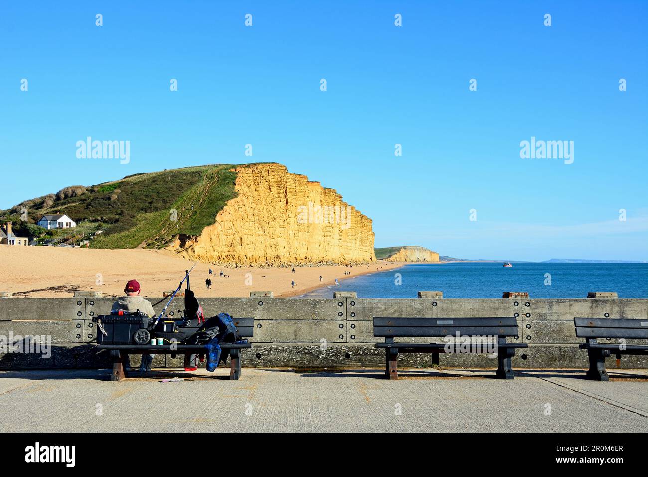 Fisherman sitting on a bench along the pier with views towards the Jurassic Coast cliffs, West Bay, Dorset, UK, Europe. Stock Photo