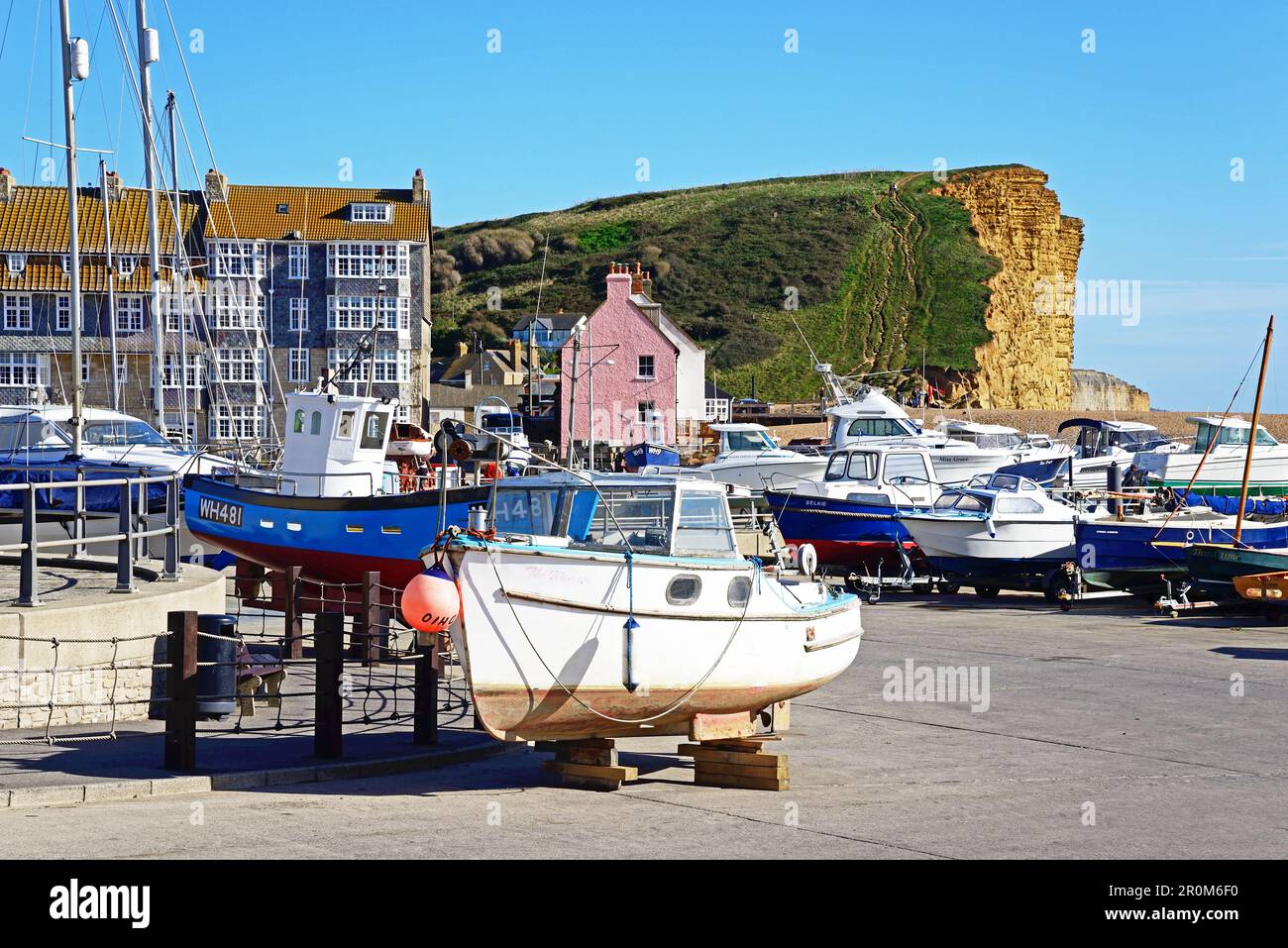 Boats in dry dock along the promenade pier with views towards the pebble beach and Jurassic Coast cliffs, West Bay, Dorset, UK. Stock Photo