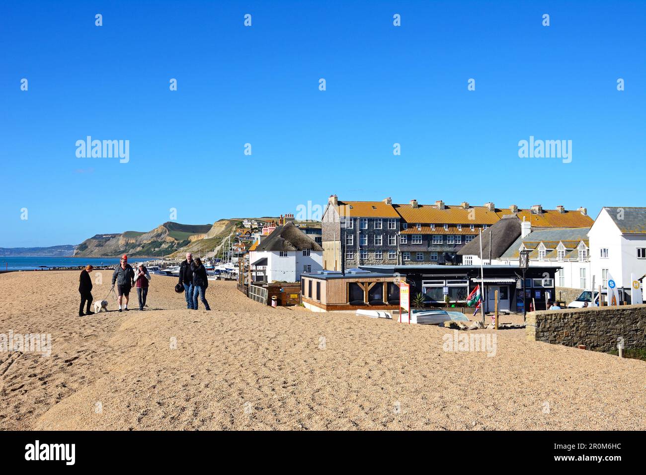 People walking along the pebble beach with views of the town and coastline, West Bay, Dorset, UK, Europe. Stock Photo