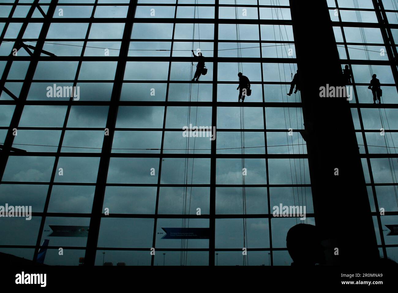 A silhouette of a worker cleaning windows hangs from a rope in the airport building. Stock Photo