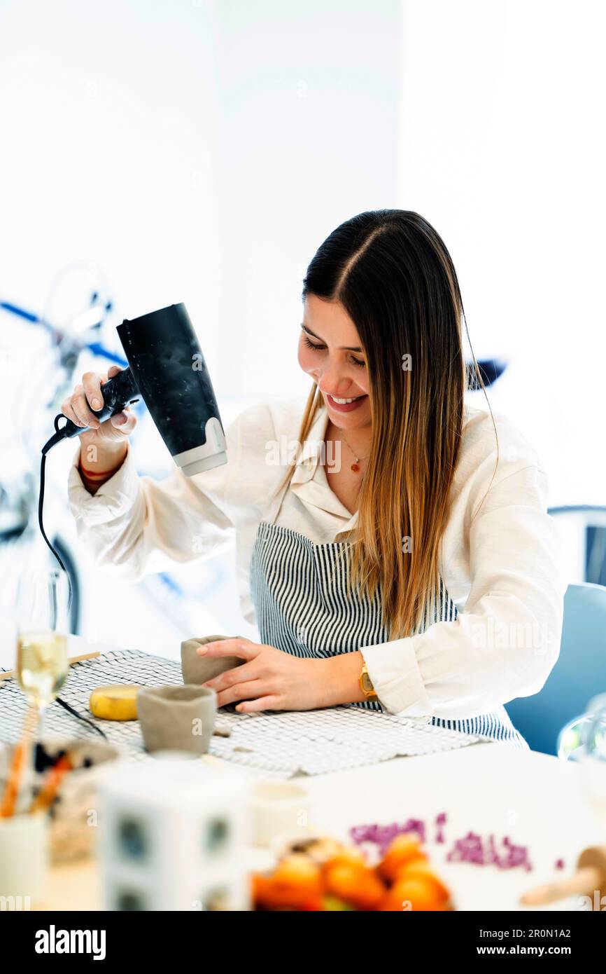 Positive young female artist in apron smiling and using dryer for clay cup while making earthenware during pottery process in studio Stock Photo