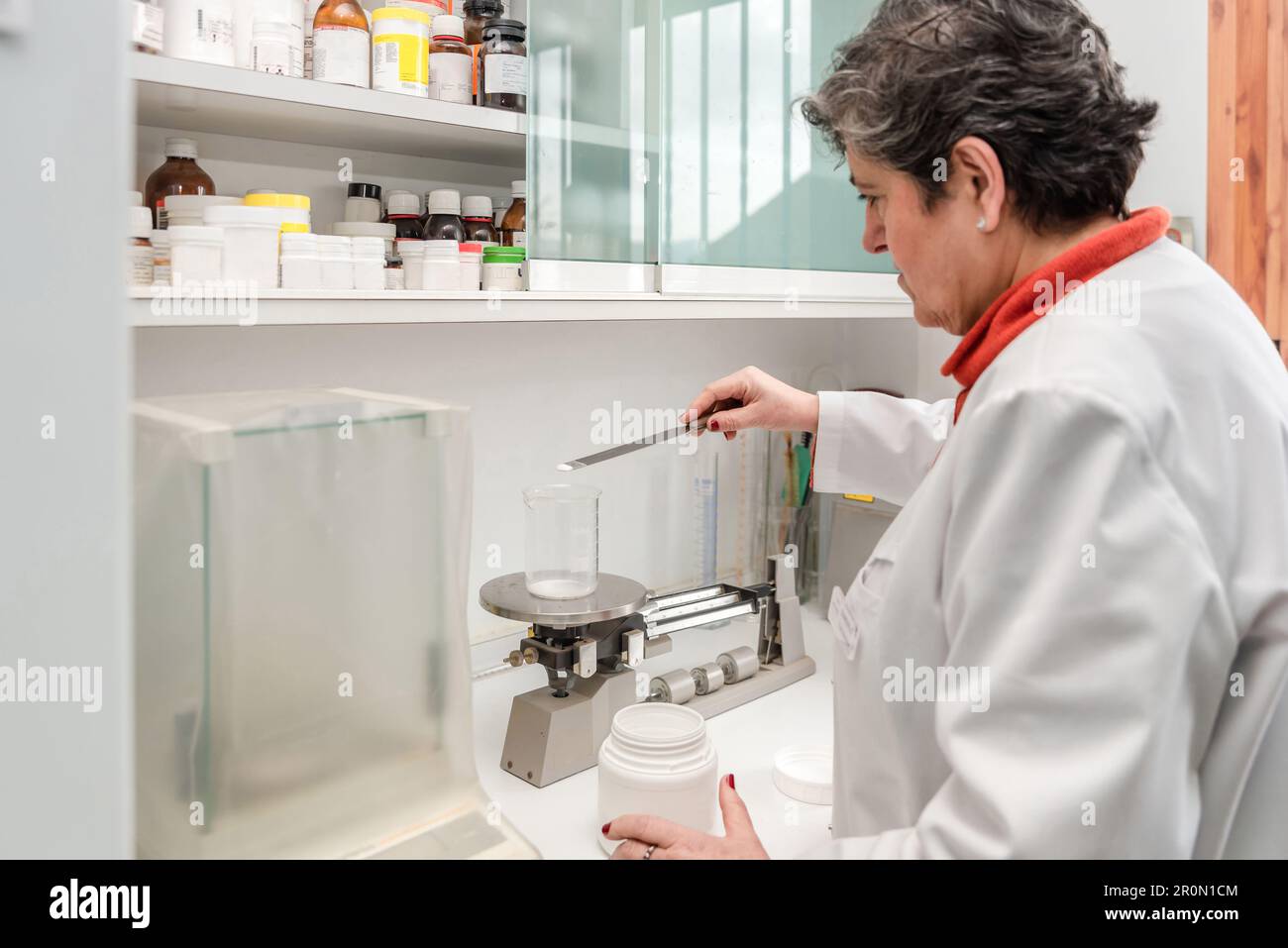 Side view of middle aged female pharmacist in white robe applying chemical pill powder on glass flask while using triple beam balance during measureme Stock Photo