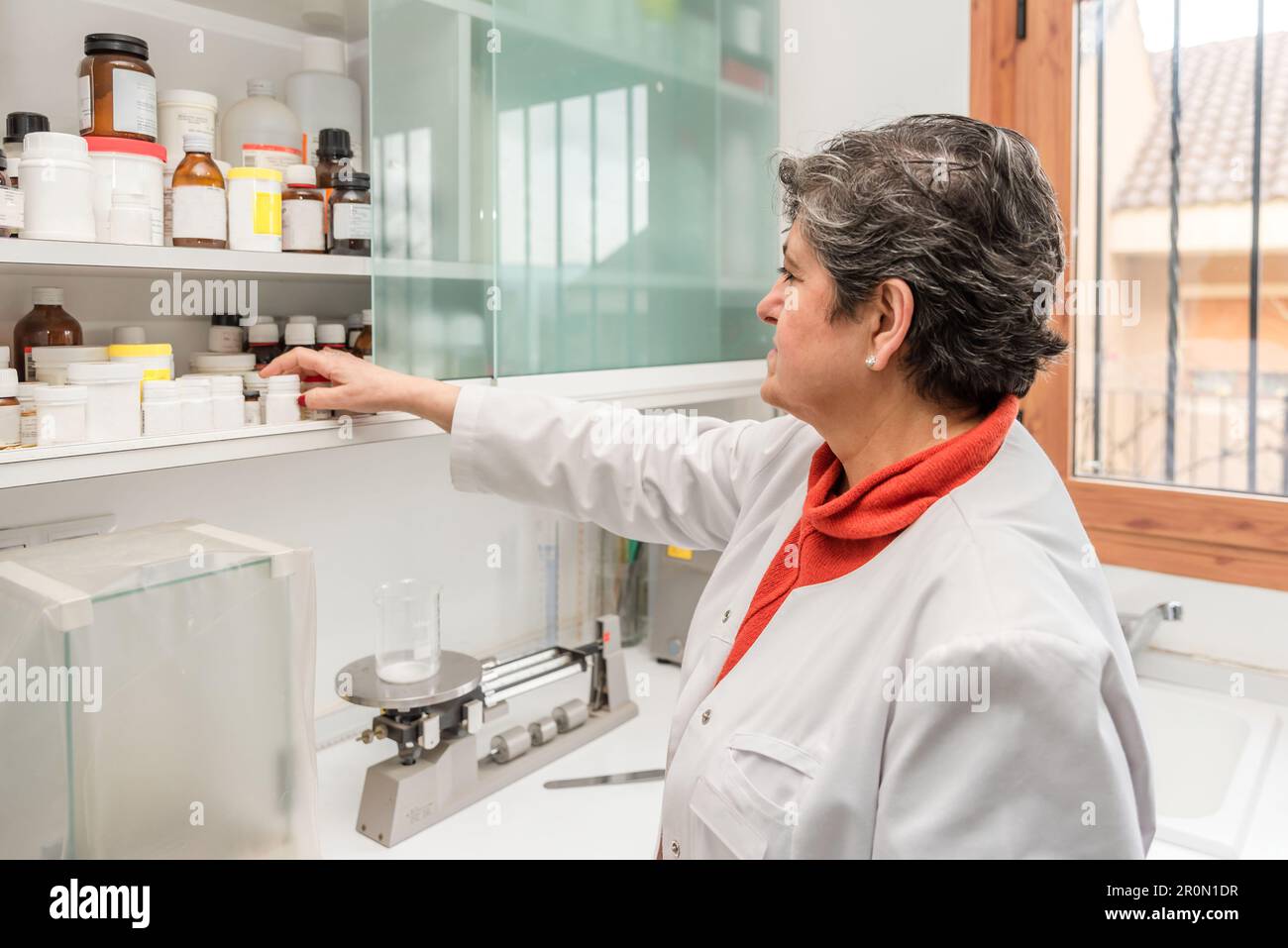 Side view of middle aged female pharmacist in medical uniform taking pills for measuring with triple beam balance instrument Stock Photo