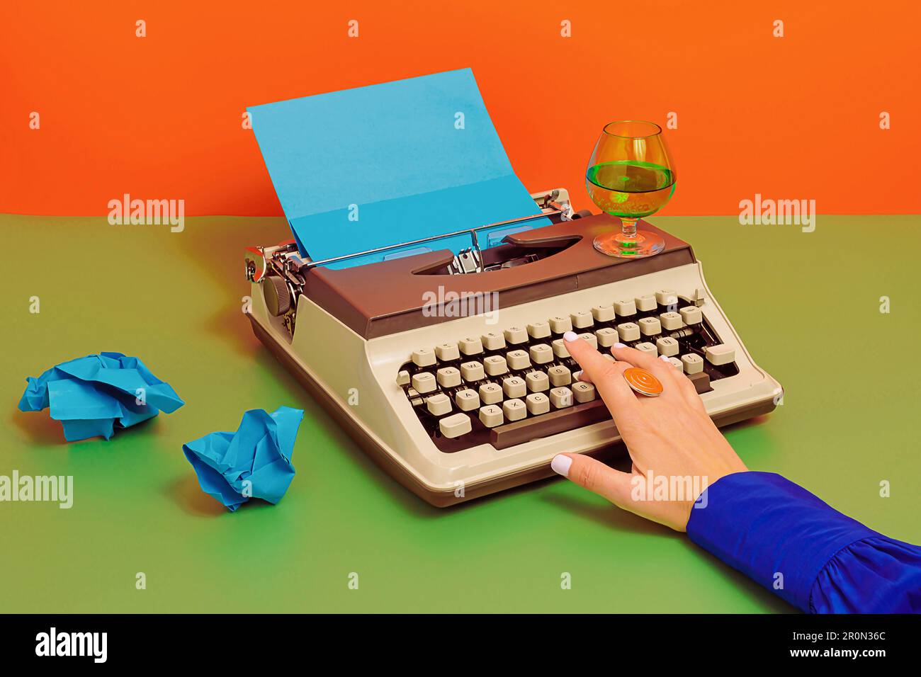 Crop anonymous person at table with glass of wine typing on old typewriter with blue carton paper over green two colored background Stock Photo