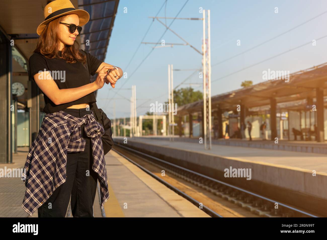 Woman in straw hat and sunglasses standing on railway station in countryside waiting train and looking at watch on her hand. Stock Photo