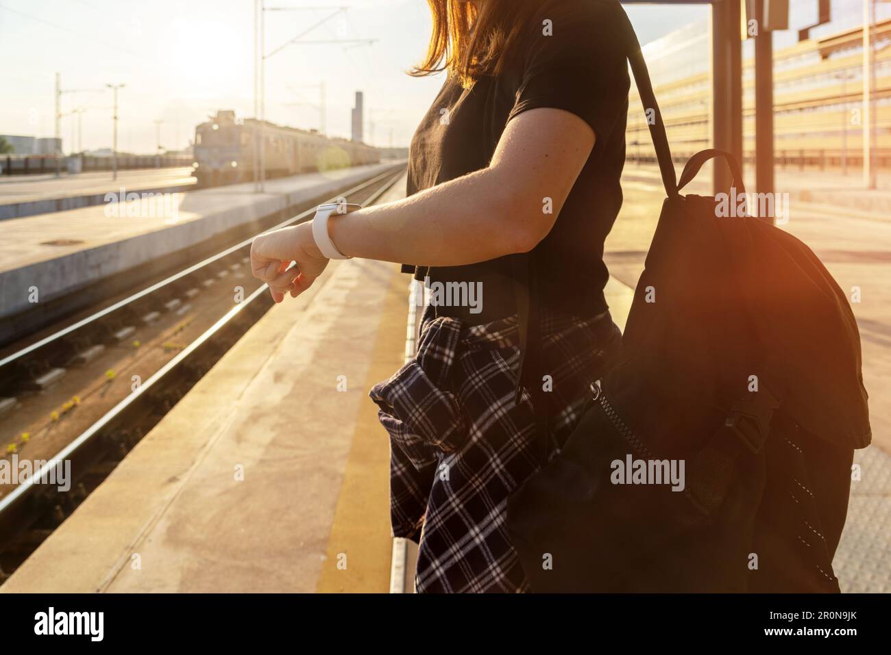 Female person standing on railway station in evening and looking at watch on her hand, train on background. Stock Photo