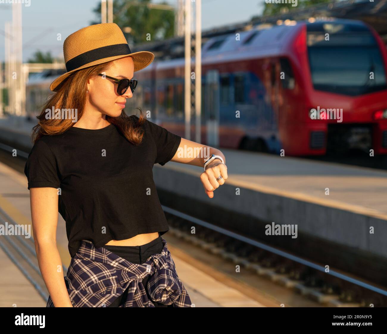 Woman in straw hat and sunglasses standing on railway platform and waiting her train, looking at watch. Stock Photo