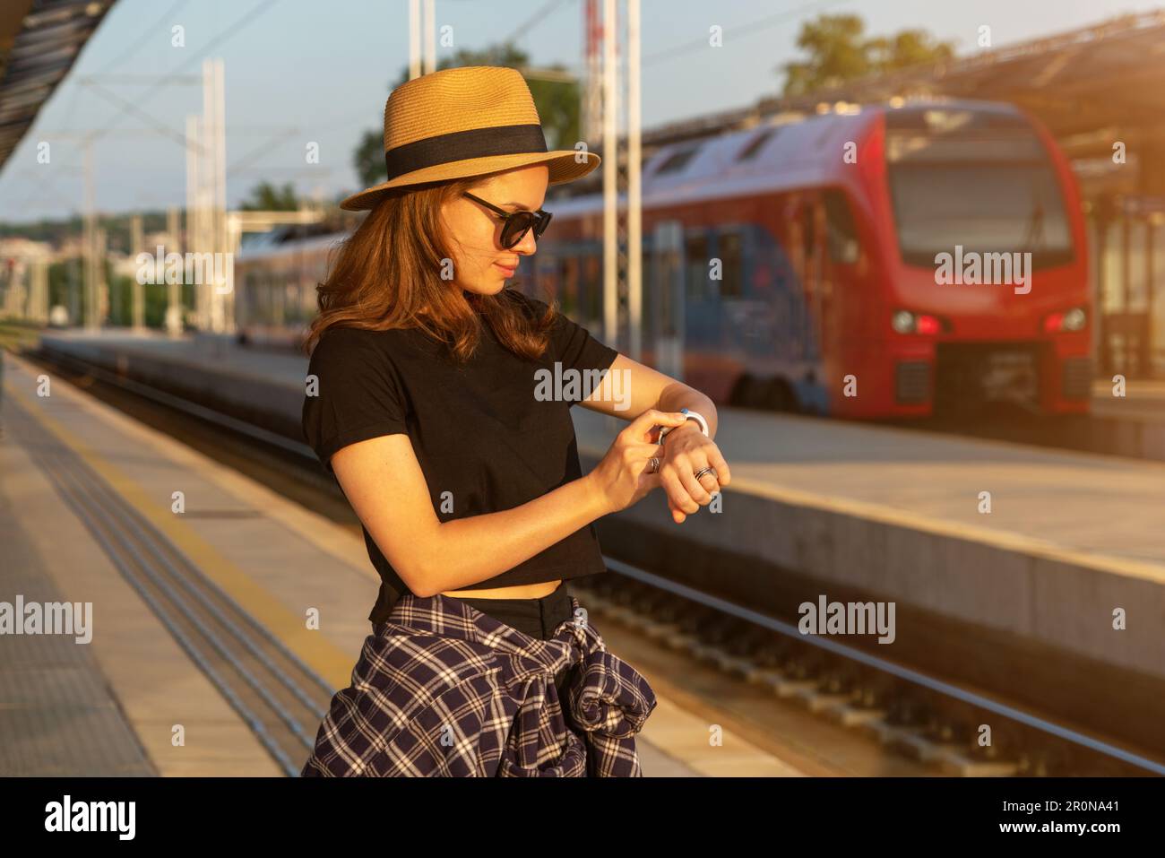 Tourist woman wearing straw hat standing on railway platform waiting train and using her smart watch. Stock Photo