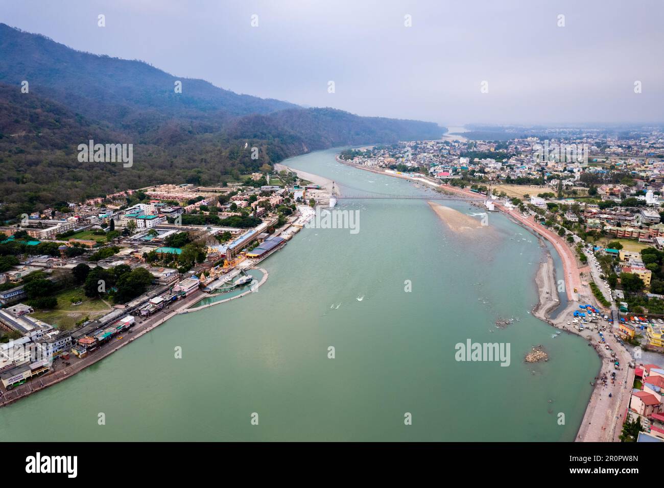 aerial drone shot of blue water of river ganga stretching into distance with himalayas with ram setu suspension bridge and temples on the banks of the Stock Photo